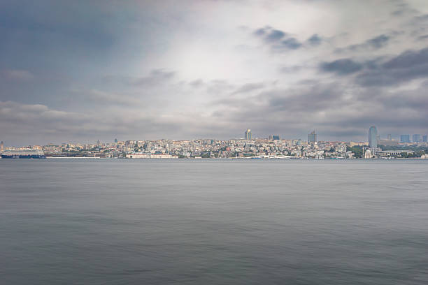 Dramatic long exposure photograph of Istanbul skyline across calm Bosphorus waters under moody overcast sky. Smooth sea surface creates serene contrast with urban cityscape.