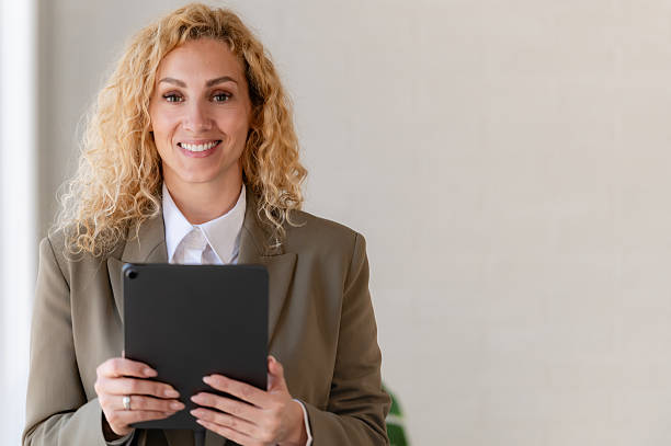 istock Smiling businesswoman using a digital tablet while standing in a bright modern office, wearing a blazer and white shirt, looking confident and professional. 2217292605