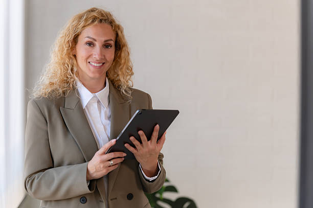 istock Smiling businesswoman using a digital tablet while standing in a bright modern office, wearing a blazer and white shirt, looking confident and professional. 2217292649