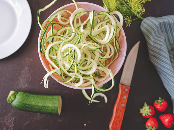 Directly above view of zucchini spaghetti in pastel pink bowl on dark brown granite kitchen counter, surrounded by dish cloth, strawberries, dill herb flowers and kitchen knife. Photo taken with Nikon Z 7II and Nikon Z 35mm f/1.8 S lens
