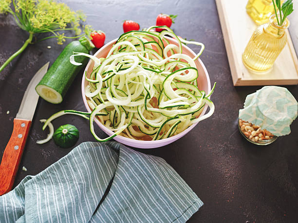 High angle view of zucchini spaghetti in pastel pink bowl on dark brown granite kitchen counter, surrounded by dish cloth, jar with pine nuts, strawberries, dill flowers and kitchen knife. Photo taken with Nikon Z 7II and Nikon Z 35mm f/1.8 S lens