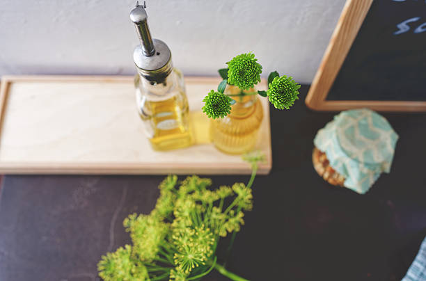 High angle view of green daisy flowers in small yellow glass bottle next to a glass bottle with olive oil on dark brown granite kitchen counter. Umbels of dill flowers and a glass jar with pine nuts next to it. Photo taken with Nikon Z 7II and Nikon Z 35mm f/1.8 S lens