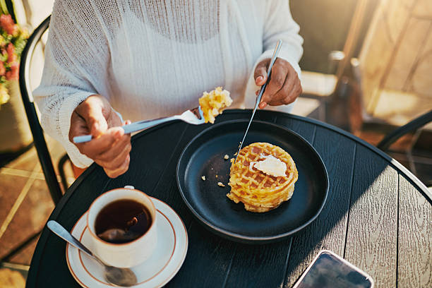 A woman in a white sweater enjoys a warm waffle topped with butter alongside a cup of tea on a sunny outdoor terrace.