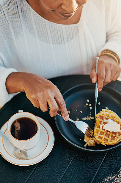 A close-up of a woman slicing a waffle on a black plate, paired with a cup of tea on a sunny outdoor table.