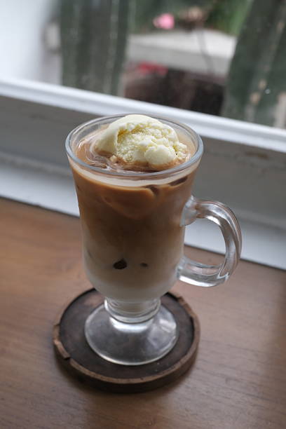 A refreshing glass of iced coffee float topped with a scoop of vanilla ice cream, served in a clear glass mug on a wooden coaster by the window