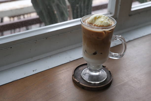 A refreshing glass of iced coffee float topped with a scoop of vanilla ice cream, served in a clear glass mug on a wooden coaster by the window