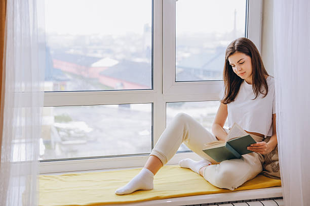 istock Young Woman Reading a Book While Relaxing by the Window at Home 2217322156