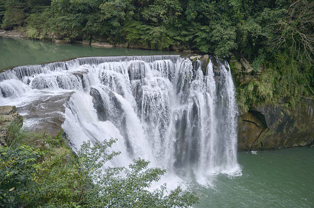 istock Powerful waterfall cascades over a wide rocky ledge into a green river pool surrounded by lush forest. 2217334042