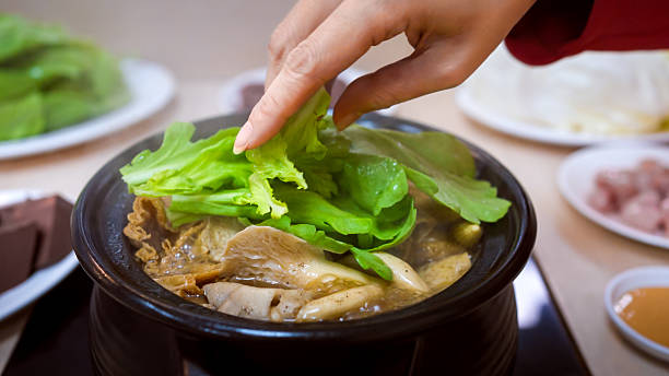Taiwanese woman eating ginger duck soup at a traditional restaurant in Taipei. She adds leafy greens to the hot soup with her hand while steam and herbs rise.