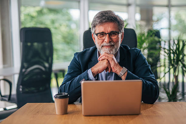 istock Senior businessman smiling at camera in modern office with laptop 2217338768