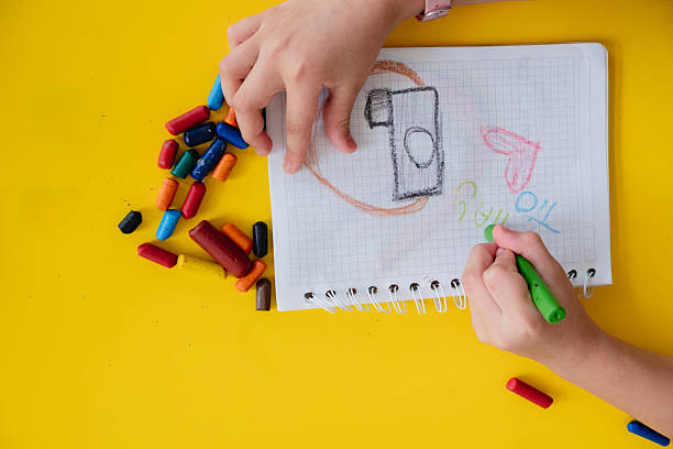 Kid drawing and playing with colorful crayons on a notepad, on a yellow background, seen from above