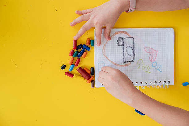 Kid drawing with colorful crayons on a notepad on a yellow surface, creating imaginative artwork
