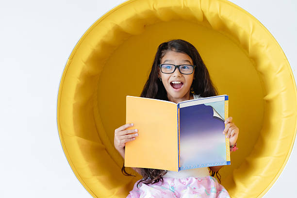 Excited elementary-aged student with glasses is sitting in a yellow chair, reading a book and looking surprised