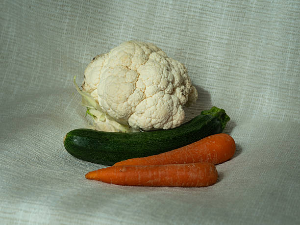 A horizontal rustic still life of fresh vegetables—orange carrots, green zucchini, and a head of cauliflower—arranged casually on softly sunlit white linen. The warm natural light highlights their organic textures and earthy tones, evoking a sense of seasonal abundance, homegrown produce and garden-fresh simplicity. A quiet homage to nature’s palette and the beauty of everyday ingredients.