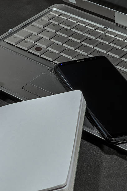 Laptop, phone, and notebook compose an office scene. The objects rest on a dark surface.