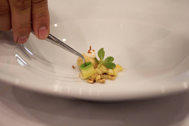 Close-up of a minimalist Mexican fine dining plate featuring toasted corn kernels, delicate zucchini rolls, verdolaga leaves, and a cooked masa ball. A chef’s hand with stainless steel tongs places the final garnish, emphasizing precision and artistry. This high-end culinary composition highlights modern Mexican cuisine’s elegance and craftsmanship, ideal for gourmet restaurant branding, culinary editorials, and fine dining promotions.