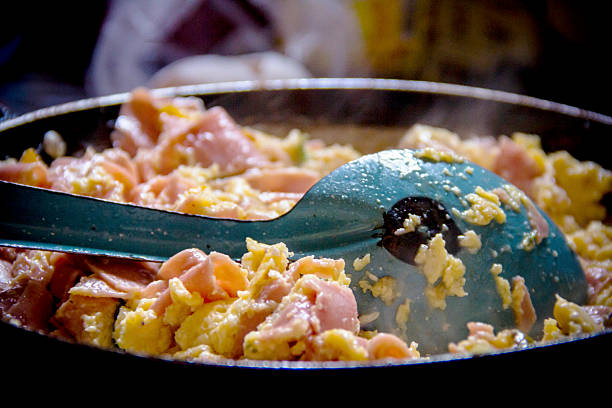 Close-up of scrambled eggs with ham cooked in a traditional blue enamelware pan, resting on a classic enamel spoon. The rustic Mexican rural setting evokes the warmth and simplicity of a daily family breakfast. Perfect for food blogs, cultural culinary projects, restaurant menus, and advertising that highlights authentic Mexican home cooking.