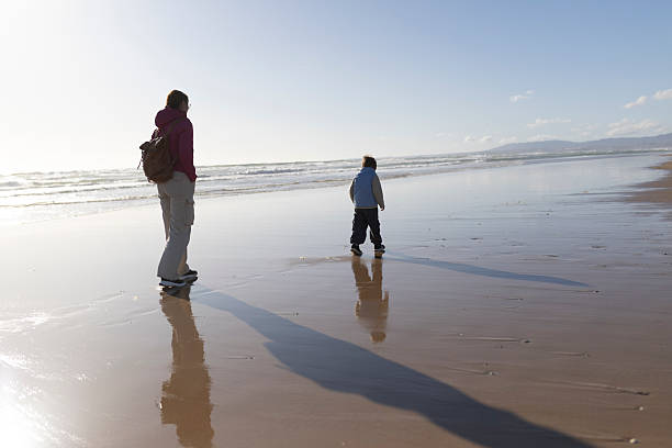 Mother and son enjoying a peaceful walk on the beach at sunset, creating long shadows on the wet sand