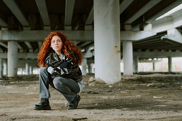 Young woman with curly red hair squatting beneath large bridge structure showcasing rugged urban surroundings feeling contemplative, wearing casual attire and black boots