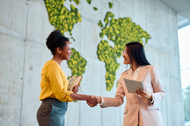Two smiling businesswomen shaking hands in modern office with moss wall map