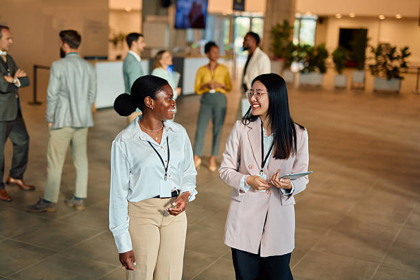 Two young businesswomen walking and talking during a break at a professional conference, smiling and enjoying their conversation while networking and connecting with each other