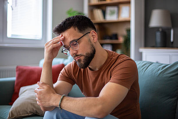 istock Depressed young man sitting on sofa touching his forehead 2217365200