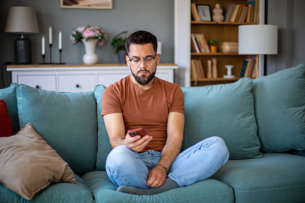 istock Young adult man sitting on sofa using smartphone at home 2217365632