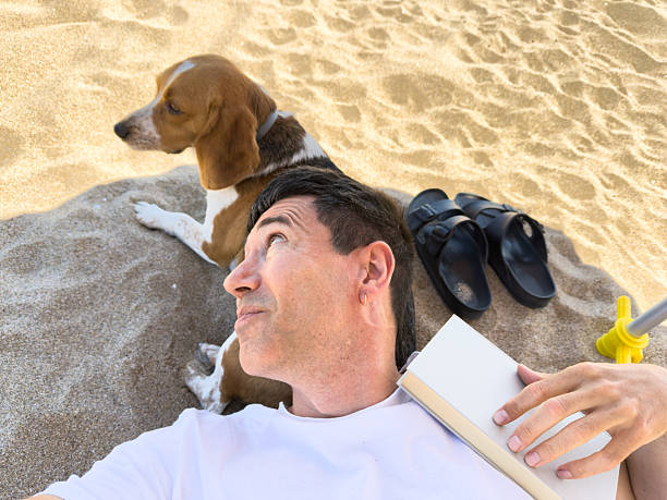 Man lying on the sand with his Beagle, holding a book and taking a selfie
