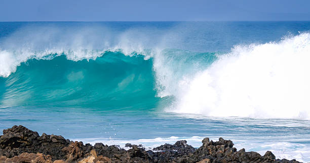 CLOSE UP: Offshore wind and powerful ocean wave breaking against rocky shore. Big wave reaches shallow waters and breaks on dark lava rocks scattered along the volcanic coastline of Canary Islands.