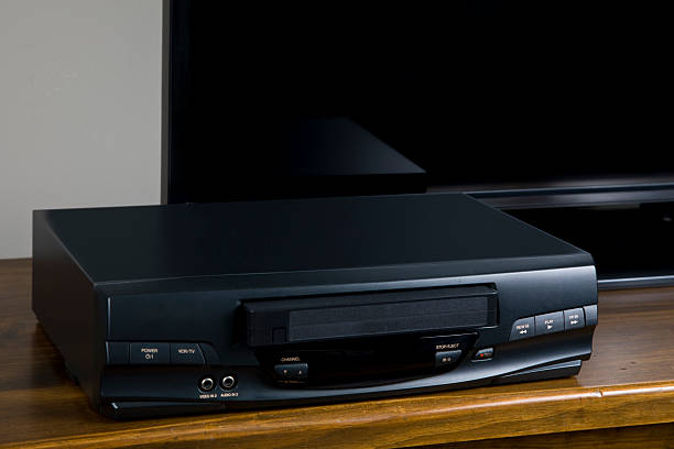 VHS video cassette player with VHS video tape inserted sitting on wooden cabinet in front of big screen TV.