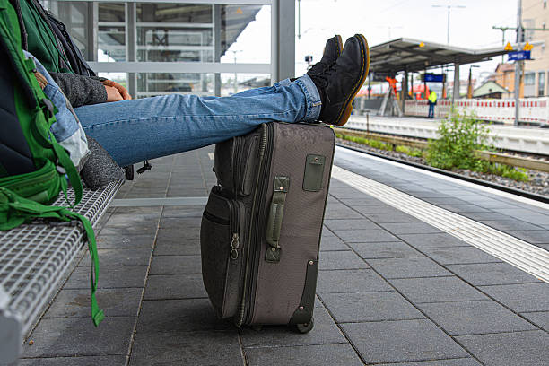 A woman is resting with legs stretched over a suitcase while waiting for a train at a railway station platform sitting on the bench surrounded with backpacks.