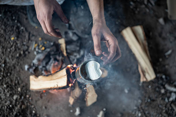 A close-up of hands holding a metal cup roasting marshmallows over a campfire. Smoke rising from the campfire provides an authentic outdoor atmosphere, evoking a sense of adventure and relaxation.
