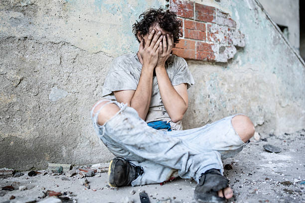 A man in distressed attire sits leaning against a weathered wall, his face hidden in despair. The setting suggests decay and abandonment, conveying emotions of struggle and vulnerability.
