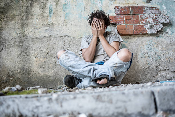 A distressed young man wearing torn clothes sits against a deteriorated wall. His hands cover his face, reflecting despair. The surroundings imply abandonment and hardship, conveying emotions of struggle and vulnerability.