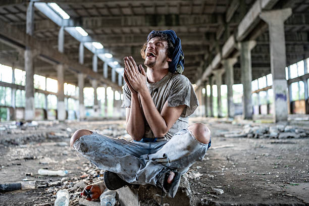 A man sits cross-legged in a dilapidated industrial space, displaying strong emotions while gesturing with clasped hands. The environment highlights a sense of abandonment and raw human resilience.