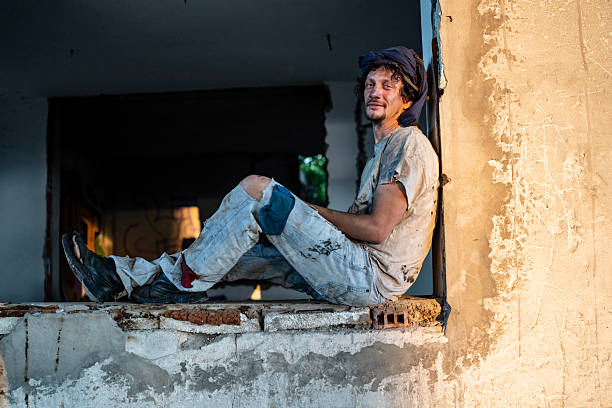 A relaxed individual in rugged clothes sits on a ledge of an abandoned building, basking in the golden light of the setting sun, surrounded by a rustic and urban atmosphere.