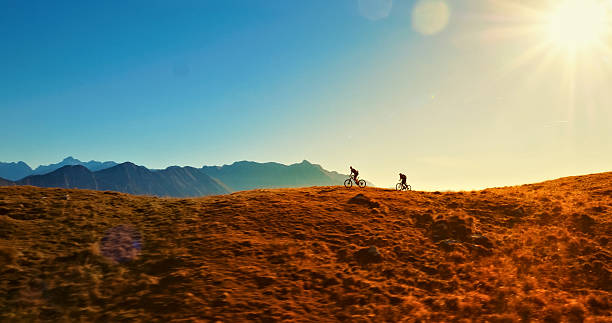 Two mountain bikers traverse a vast, grassy hilltop as the sun sets, casting a warm glow over the landscape. The aerial view highlights the thrill of outdoor adventure and the beauty of nature.