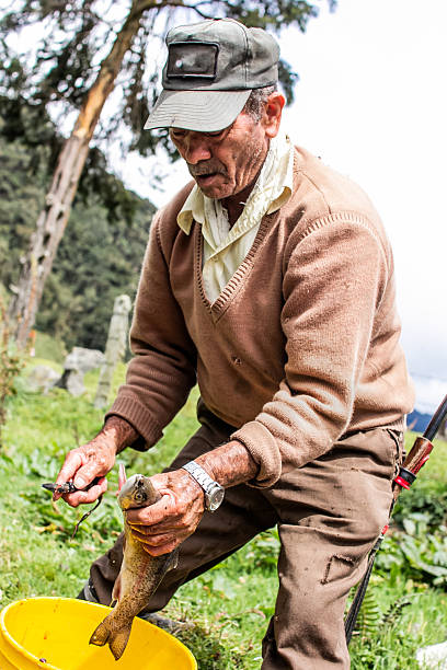 An older farmer proudly holds a freshly caught trout as he cleans it next to a yellow bucket in a mountainous area. The image reflects rural work, artisanal fishing and deep connection with nature