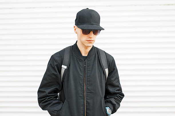 istock Young handsome guy in a black jacket with a baseball cap and a backpack near a white metal wall 2217379911