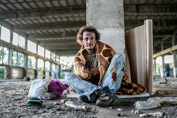 A man sits on the floor of an abandoned building, surrounded by personal belongings and rubble. The image conveys hardship, resilience, and the stark reality of homelessness in an urban setting.
