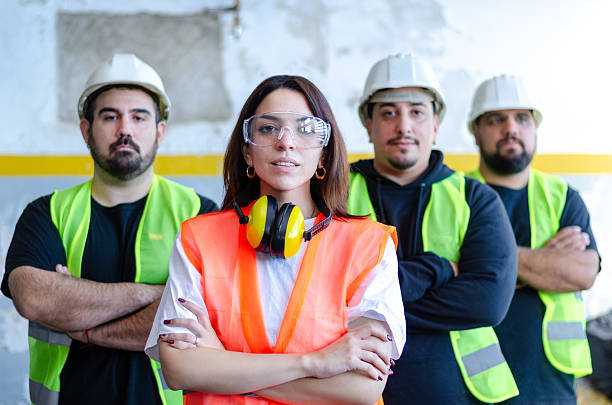 istock Confident young female engineer wearing a hard hat and reflective vest stands with arms crossed at a construction site, with colleagues in the background. 2217386364