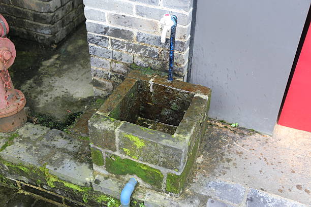 A small square brick well with moss growing on the bricks. There is an old blue water pipe connected to it, and green pipes next to them, located in front of gray walls with red doors