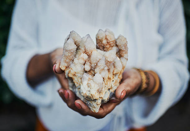 A close-up of feminine hands holding a sparkling quartz crystal cluster with earthy tones, set against a blurred natural background, evoking organic beauty.