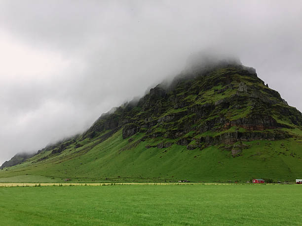 An iconic volcanic mountain in southern Iceland, partially obscured by low-hanging clouds. The steep, jagged rock face is covered with vibrant green moss, contrasting sharply with the overcast sky. Shot on iPhone.