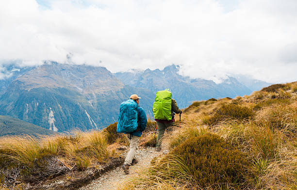 Rear view of two backpackers hiking through wild trail holding hiking poles, A day out in nature, Routeburn Track, South Island, New Zealand.