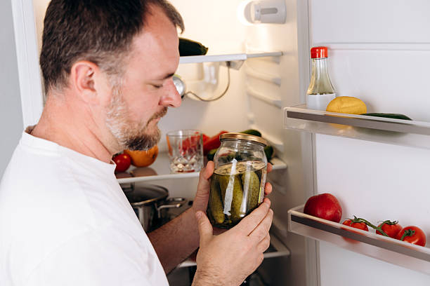 istock Man inspecting jar of pickles while standing in front of open refrigerator filled with fresh vegetables and condiments, showcasing food preservation and healthy eating habits in a modern kitchen environment 2217417668