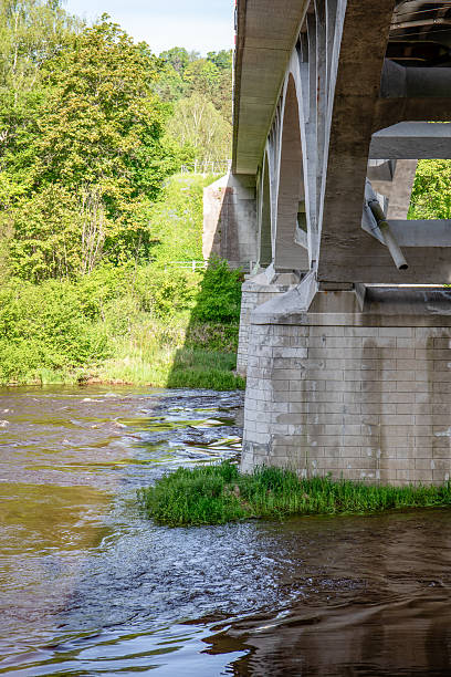 istock Concrete Bridge Structure Over Flowing River with Green Trees and Reflections in Daylight 2217423119