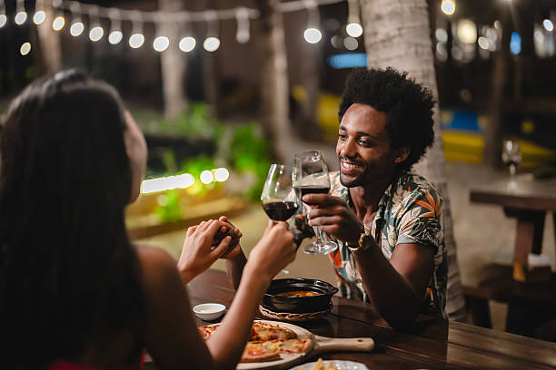 Romantic couple enjoying an intimate dinner under string lights at night, holding hands and smiling while dining outdoors at a tropical beachside restaurant.