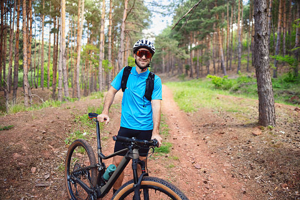 A young male cyclist stands confidently in a lush forest, holding his mountain bike. The trail is surrounded by vibrant greenery, ideal for an adventurous outdoor experience resembling nature and freedom.