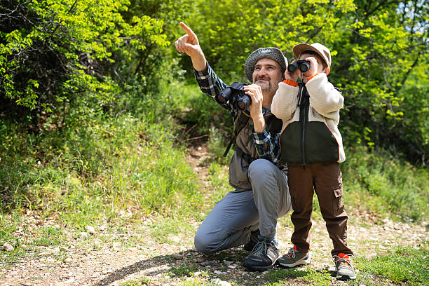 A father and his young son explore nature together, observing birds with binoculars in a verdant forest setting filled with greenery and sunlight, promoting bonding and appreciation for wildlife outdoors.